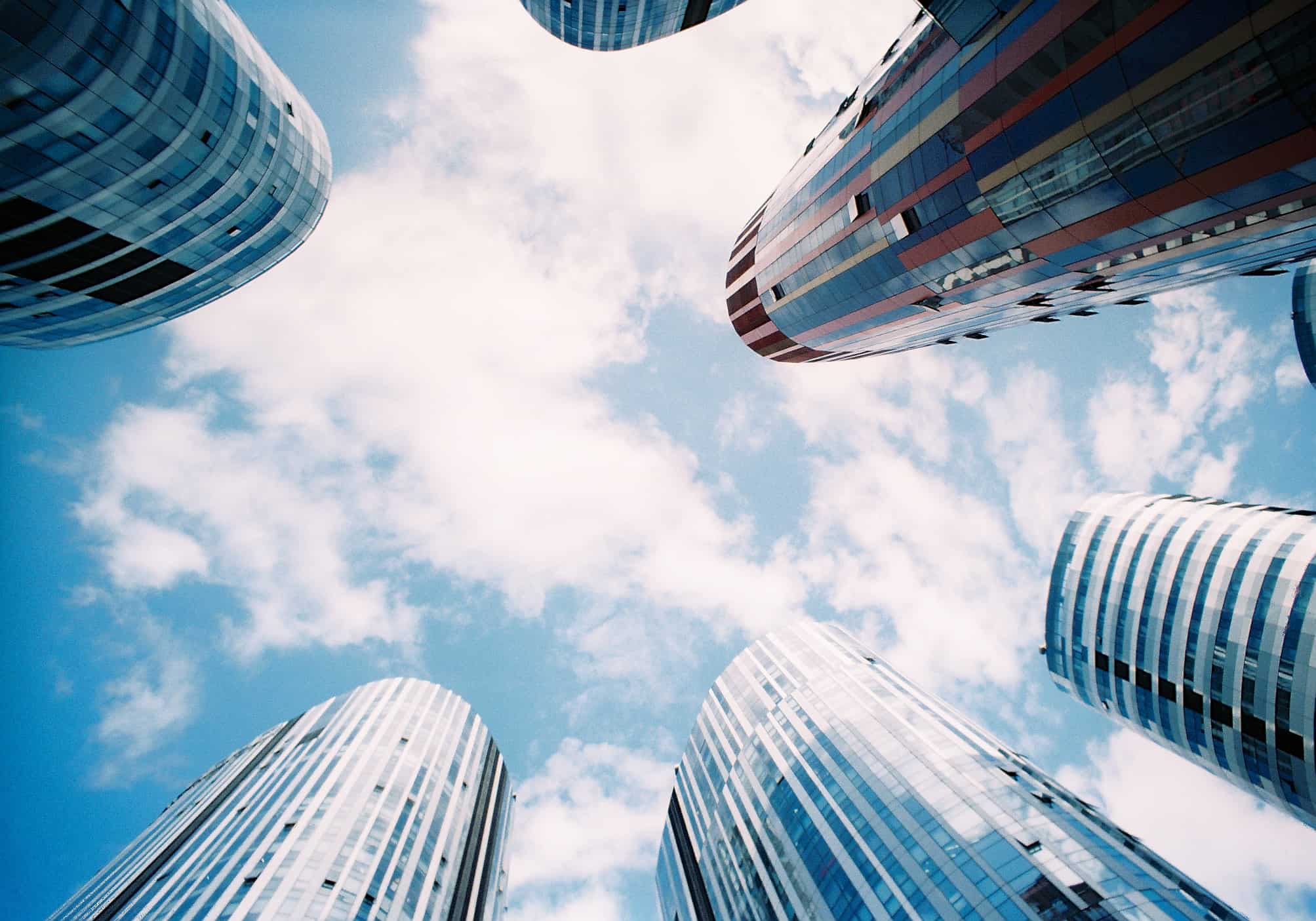 Skyscrapers with reflective glass facades curve inward around the edges of the image, looking up toward a blue sky with scattered clouds—a cityscape echoing the surreal rhythm of glossolalia.
