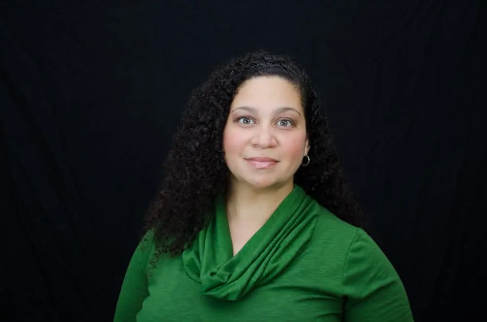 A woman with curly dark hair wearing a green top is posing in front of a plain black background, looking directly at the camera with a neutral expression.