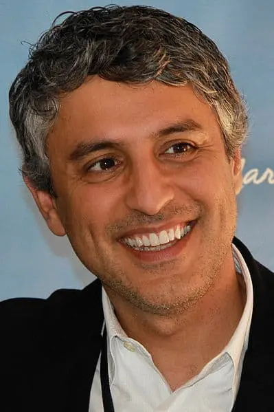 A man with short, wavy salt-and-pepper hair smiles warmly. He is wearing a white collared shirt under a dark jacket, standing in front of a blurry blue background.