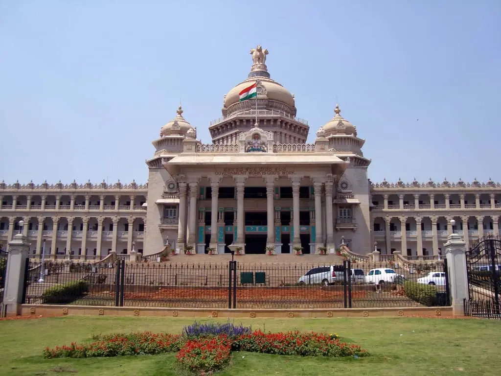 A large, ornate government building with domes and columns, featuring the Indian flag on top. A manicured lawn and iron fence are in the foreground under a clear sky.
