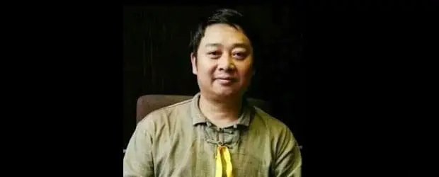 A man with short dark hair wearing a light brown shirt sits and faces the camera, smiling slightly. He has a yellow ribbon around his neck, a subtle nod to ongoing discussions about censorship in China. The background is dark.
