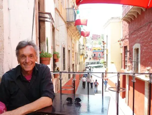 A smiling man sits at an outdoor table on a balcony overlooking a colorful street with flags, potted plants, and people walking below on a sunny day.
