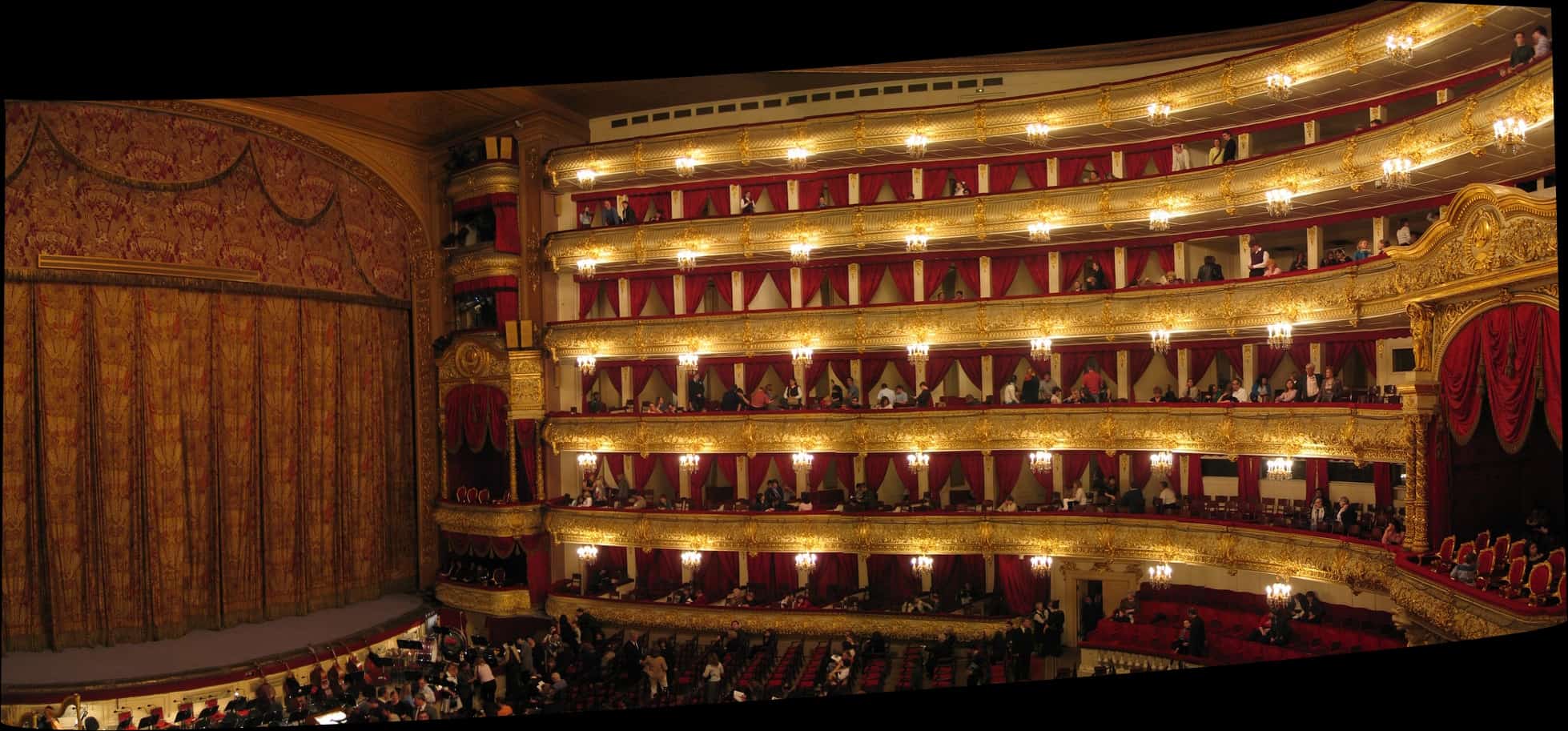 A grand theater interior with ornate balconies, red velvet curtains, and gold detailing. The audience is seated, and the stage curtain is closed, with warm lighting illuminating the space.