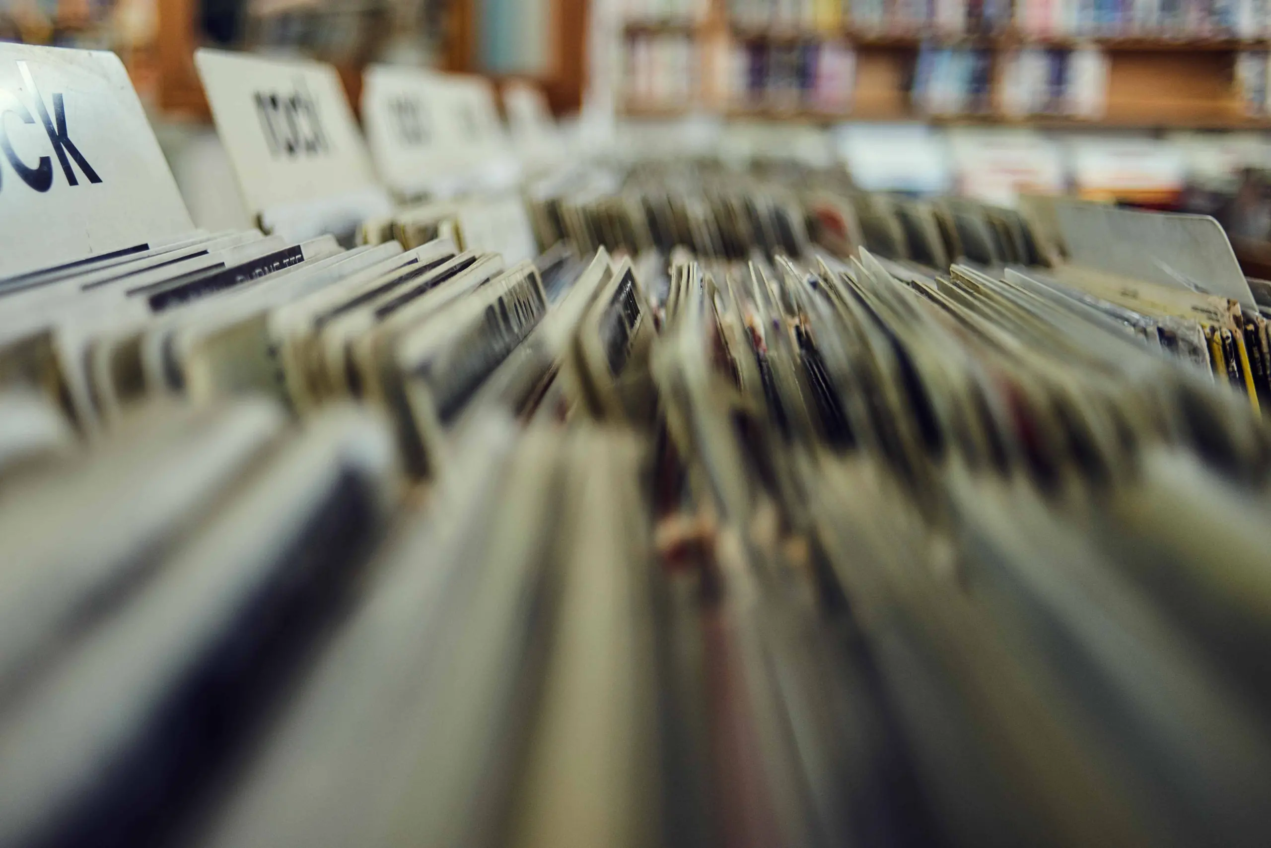 Close-up of rows of vinyl records in a record store, with category dividers labeled ROCK and JOSH. The image is focused on the records in front, with a blurred background of more shelves.