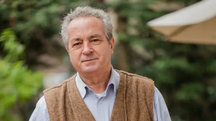 An older man with curly gray hair, wearing a light blue shirt and a brown vest, stands outdoors with blurred greenery and an umbrella in the background.