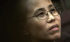 A person with short hair and glasses looks slightly upward, appearing thoughtful. The background is out of focus, emphasizing their face.