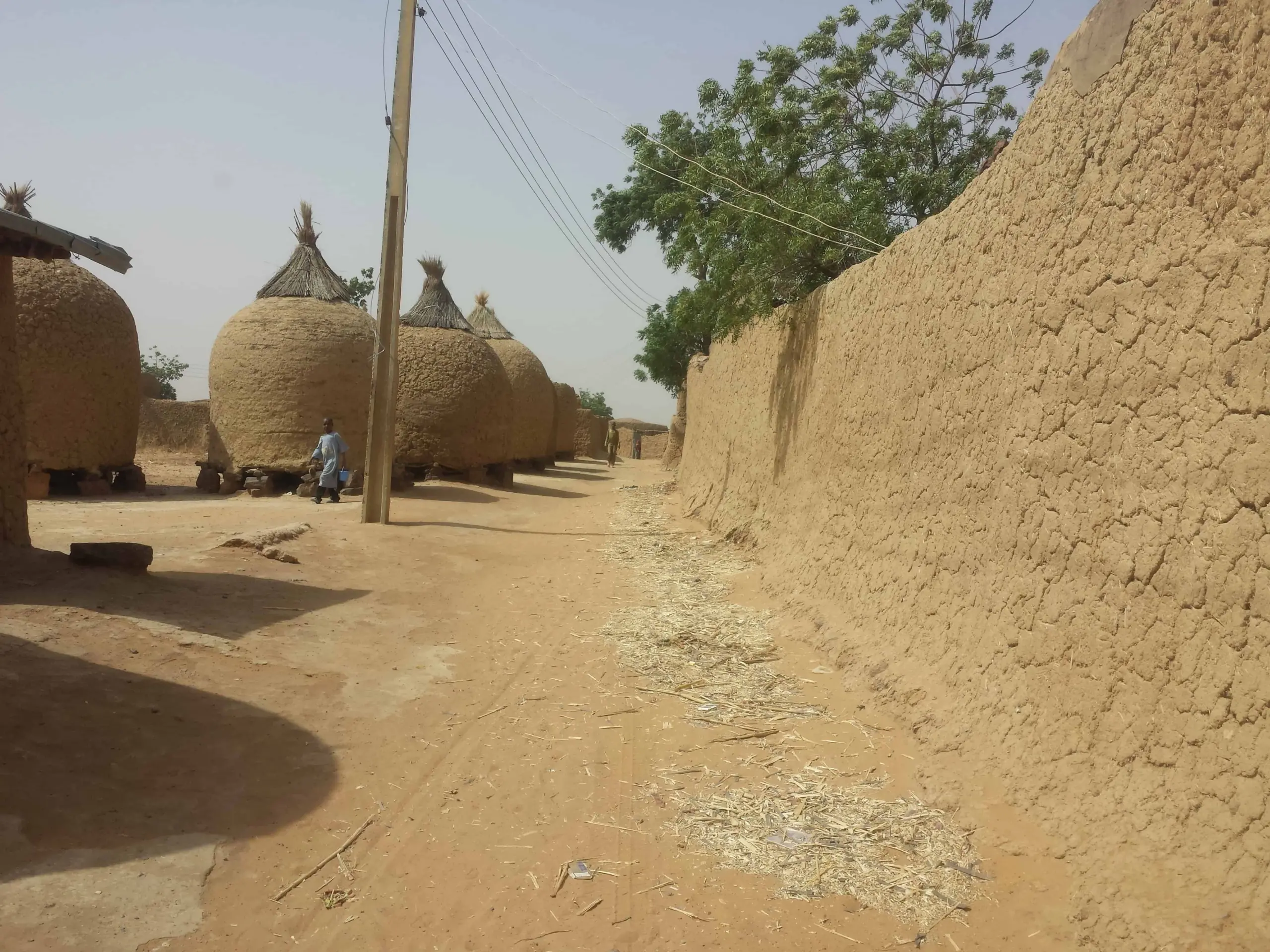 A sandy street lined with large, round, thatched-roof mud huts and high mud walls, with a person walking in the background under clear skies. Electric wires run along wooden poles beside the huts.