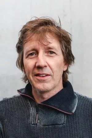 A middle-aged man with tousled brown hair and blue eyes, wearing a dark zip-up sweater, stands in front of a plain light-colored background, looking directly at the camera with a neutral expression.