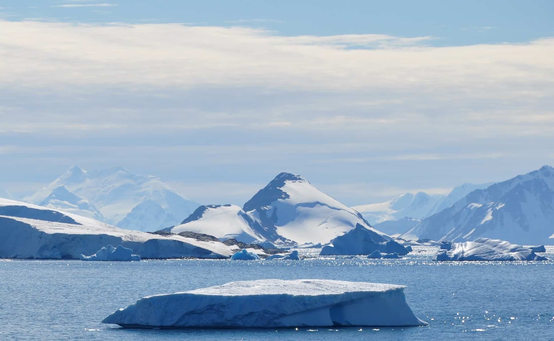 A floating iceberg drifts in calm blue water near Heim, with snow-covered mountains and glaciers in the background under a bright, partly cloudy sky.