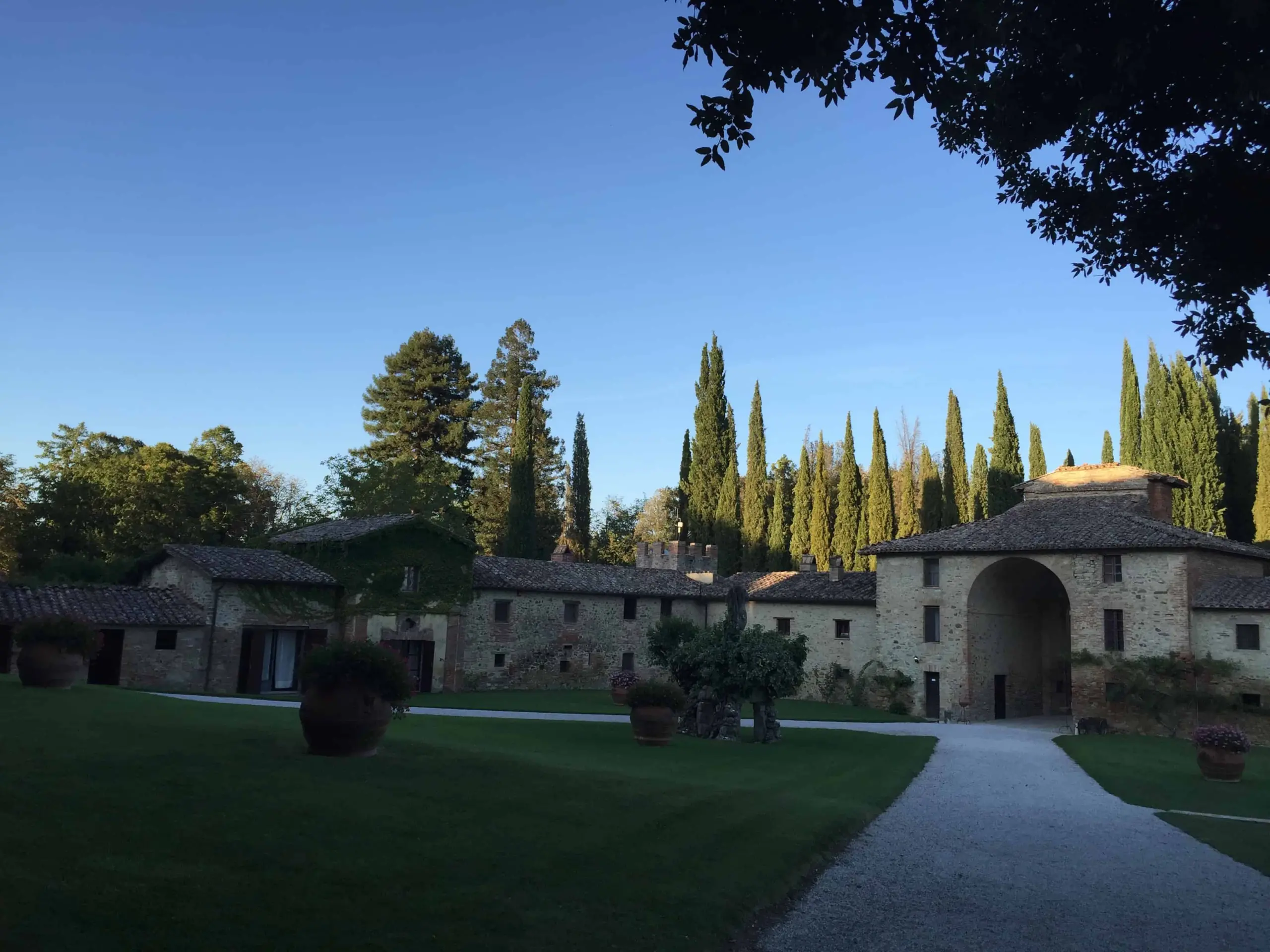 A large stone villa with arched entryways and tiled roofs is surrounded by tall cypress trees, green lawns, and potted plants under a clear blue sky.