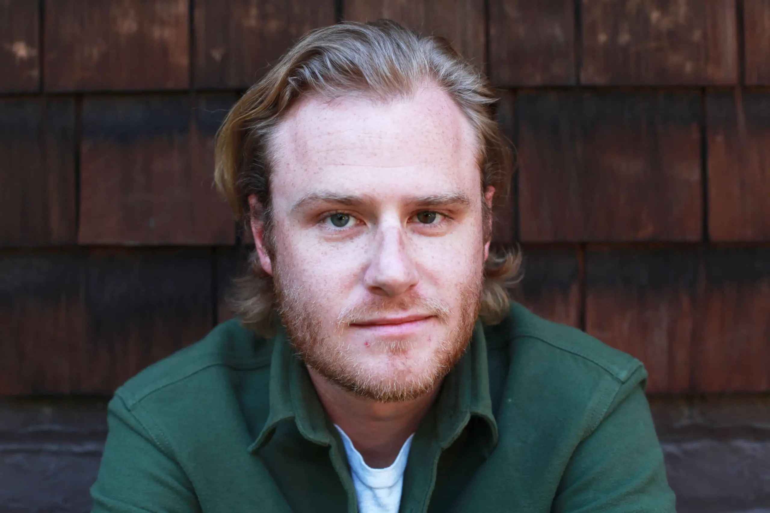 A man with light skin, medium-length blond hair, and a short beard wears a green jacket and white shirt, sitting in front of a wooden shingle wall, looking directly at the camera with a neutral expression.