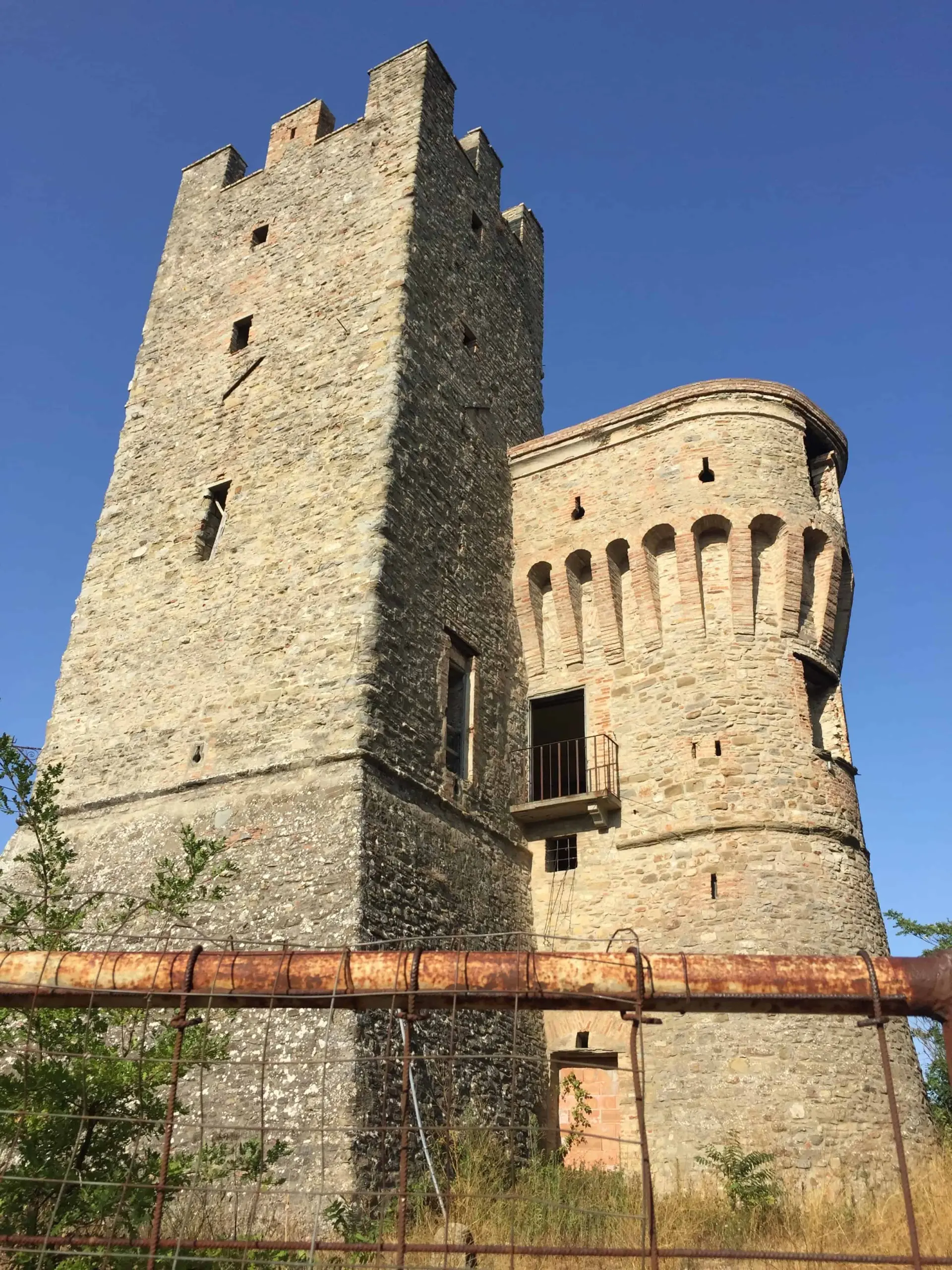 A stone medieval tower and fortress with narrow windows and crenellations, seen from below against a clear blue sky, with a rusted metal fence and some green shrubs in the foreground.