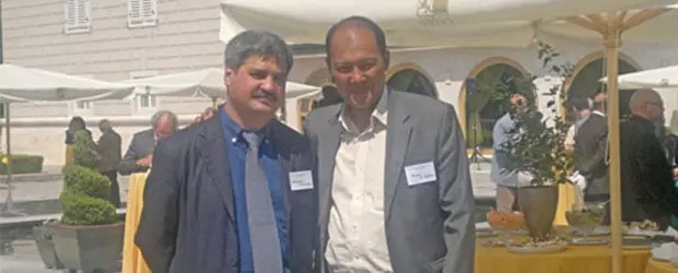 Two men in suits stand close together outdoors at a social event with tables and yellow tablecloths, surrounded by other people and umbrellas, in front of a light-colored building.