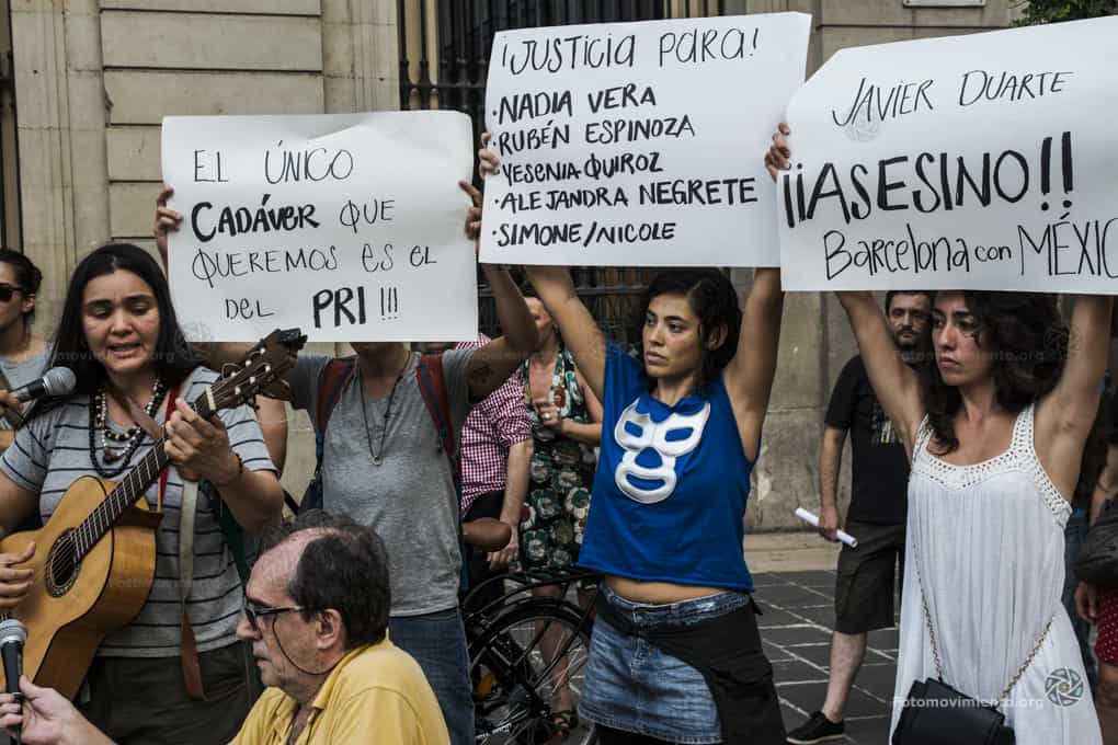 A group of people protest outdoors. Three women hold signs in Spanish demanding justice for victims and accusing Javier Duarte. One woman plays guitar and sings, while others stand nearby, one seated in front.