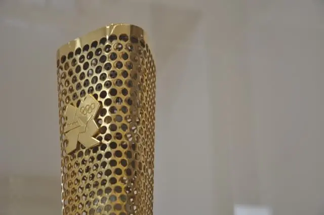 A close-up of the gold London 2012 Olympic torch, its perforated design with circular holes and the Olympic logo engraved on its surface, evokes the spirit of resilience seen in figures like Liu Xiaobo.