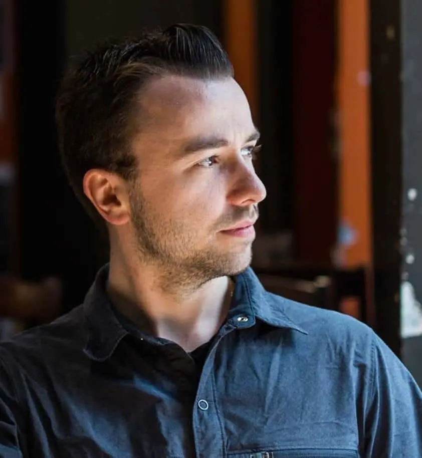 A man with short dark hair and light stubble, wearing a blue button-up shirt, looks thoughtfully to his left. The background is softly lit with dark and orange tones.