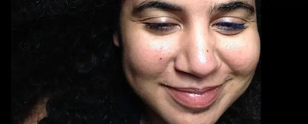 A person with curly dark hair smiles with their eyes closed, showing dimples and a natural, relaxed expression. The background is dark, highlighting their face.