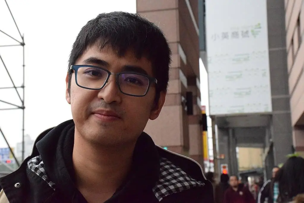 A young man with short dark hair and glasses stands outdoors in an urban setting, wearing a jacket and a black hoodie. Buildings and a blurry sign in Chinese are visible in the background.