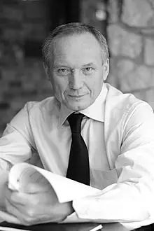 A middle-aged man in a dress shirt and tie sits at a table, holding a rolled-up document and looking directly at the camera with a slight smile. The background is softly blurred with brick and stone.