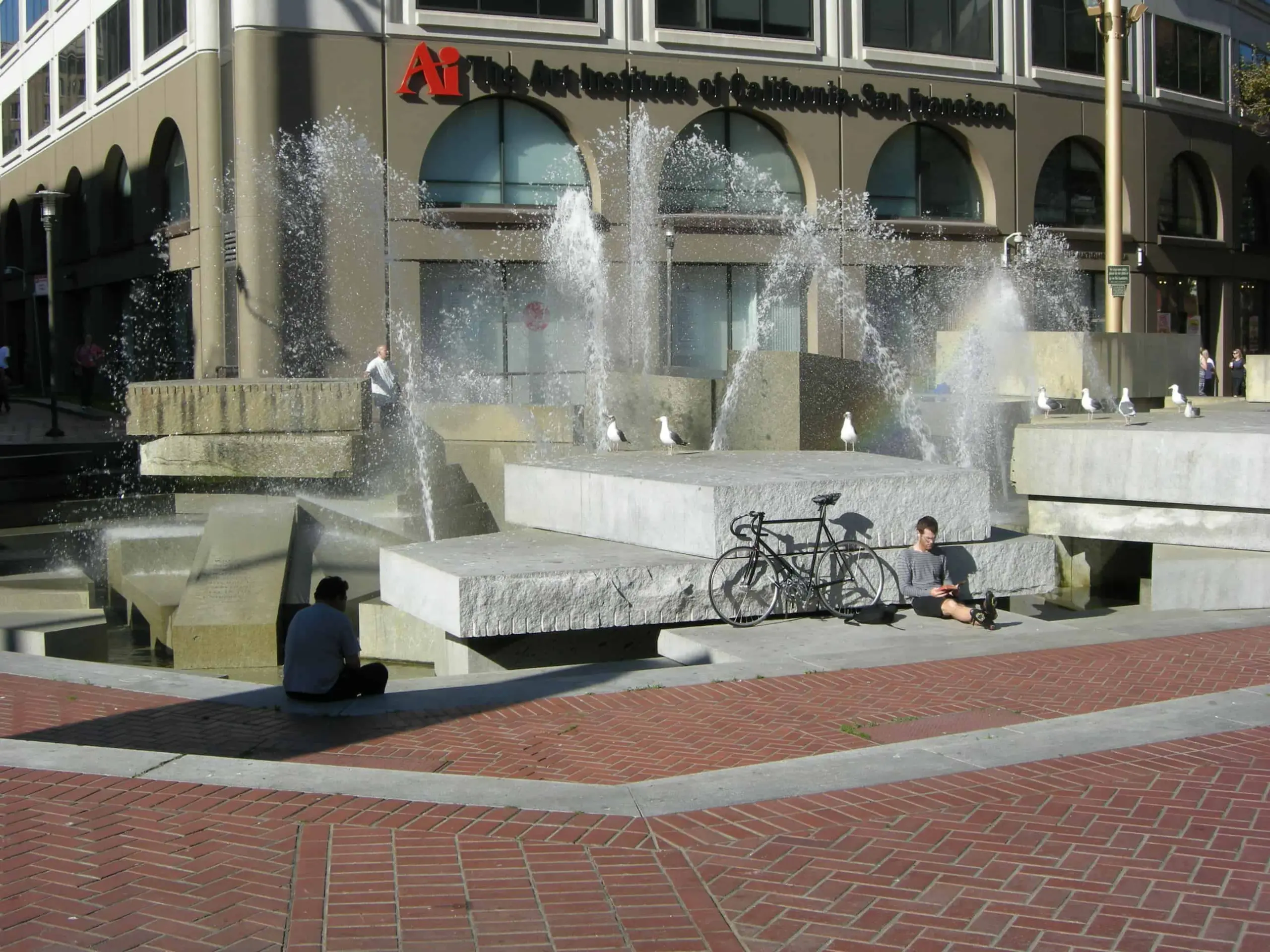 A city plaza with a modern fountain featuring multiple streams of water. Two people sit on the edge near a bicycle. The Art Institute of California–San Francisco building is in the background.