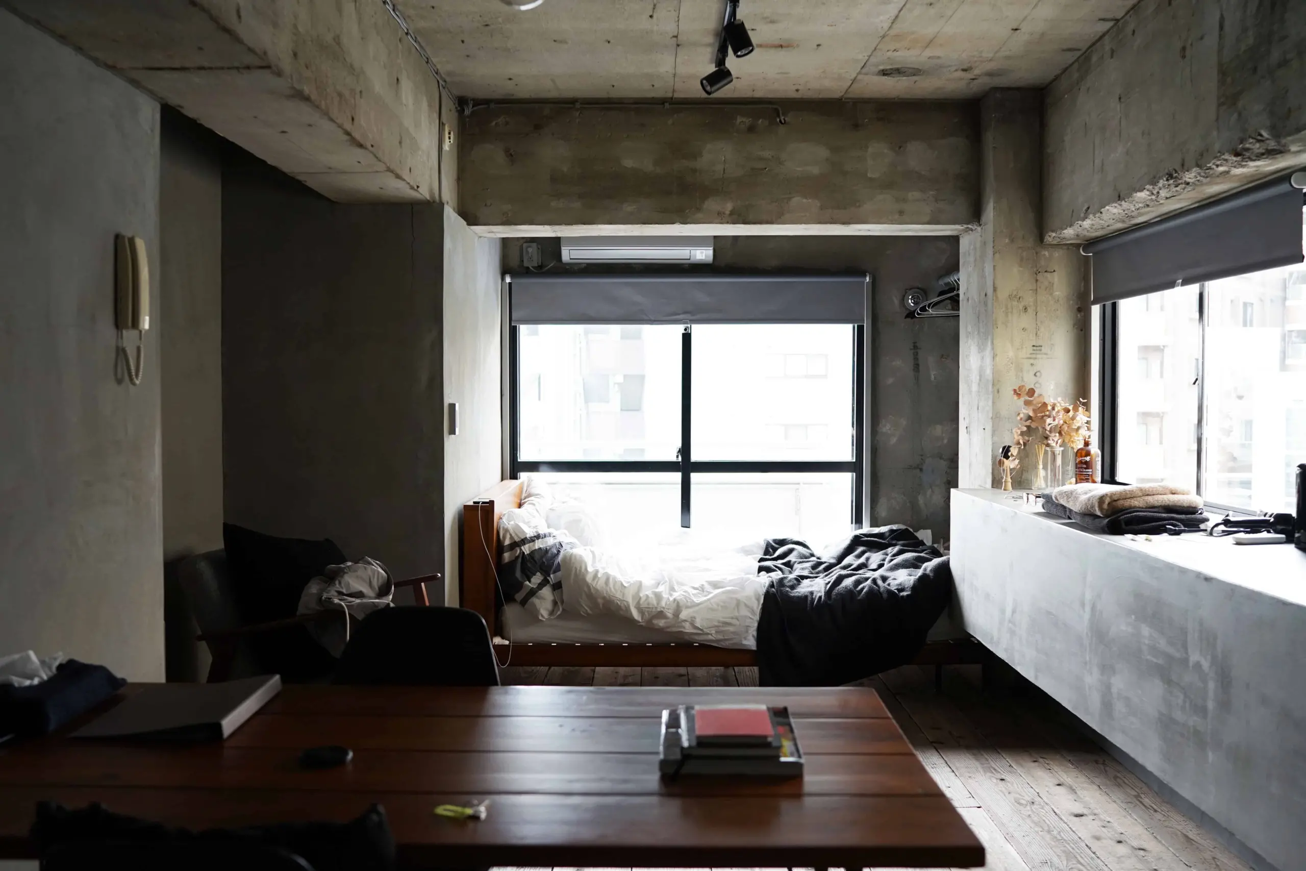 A minimalist, industrial-style bedroom with concrete walls and ceiling, a large window behind an unmade bed with white and black bedding, a wooden table with books, and dried flowers on a side counter.