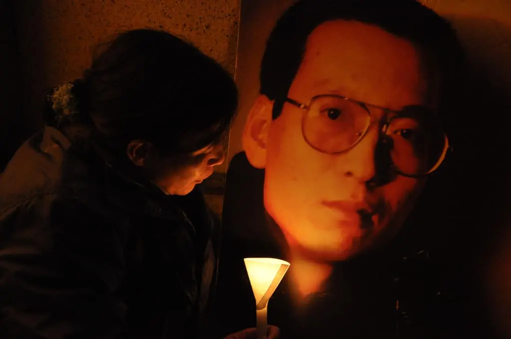 A person holding a lit candle stands beside a large portrait of a man wearing glasses, illuminated by the candlelight in a dark setting, suggesting a vigil or memorial.