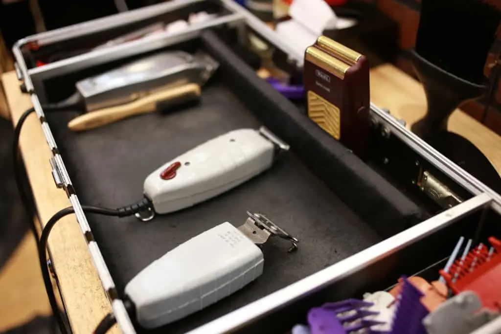 A close-up of a barber’s tool case, evoking the precision seen in prison writing, contains electric hair clippers, a brush, a razor, and various comb attachments—all neatly organized inside a black tray.