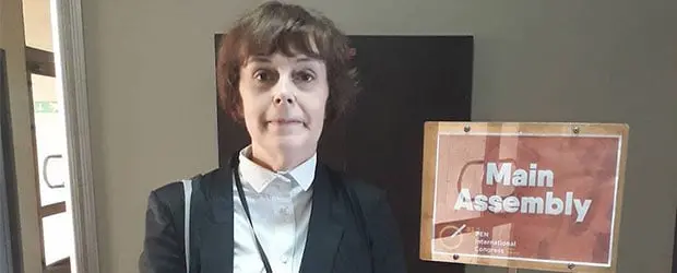 A person with short brown hair, wearing a black blazer and white shirt, stands next to a sign that reads Main Assembly indoors.