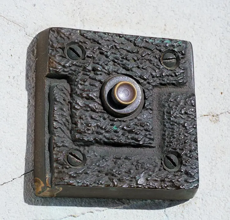A close-up of a textured, metal doorbell button mounted on a wall, secured by four screws at each corner. The rough, hammered appearance evokes the rawness found in prison writing.