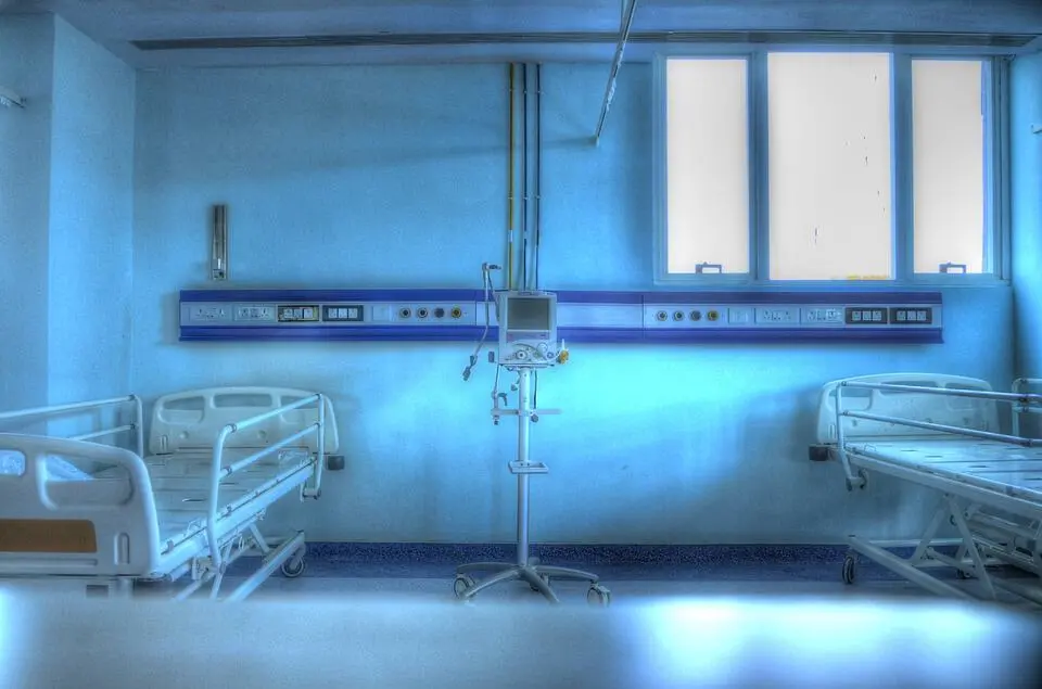Two empty hospital beds with white frames are positioned on either side of a medical monitor in a brightly lit, blue-toned hospital room, their stillness echoing the isolation often described in prison writing. Large windows are visible in the background.