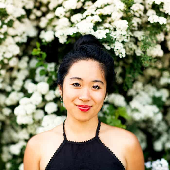 A woman with dark hair in a bun, wearing a black halter top and red lipstick, stands in front of a background filled with blooming white flowers. She is smiling slightly at the camera.