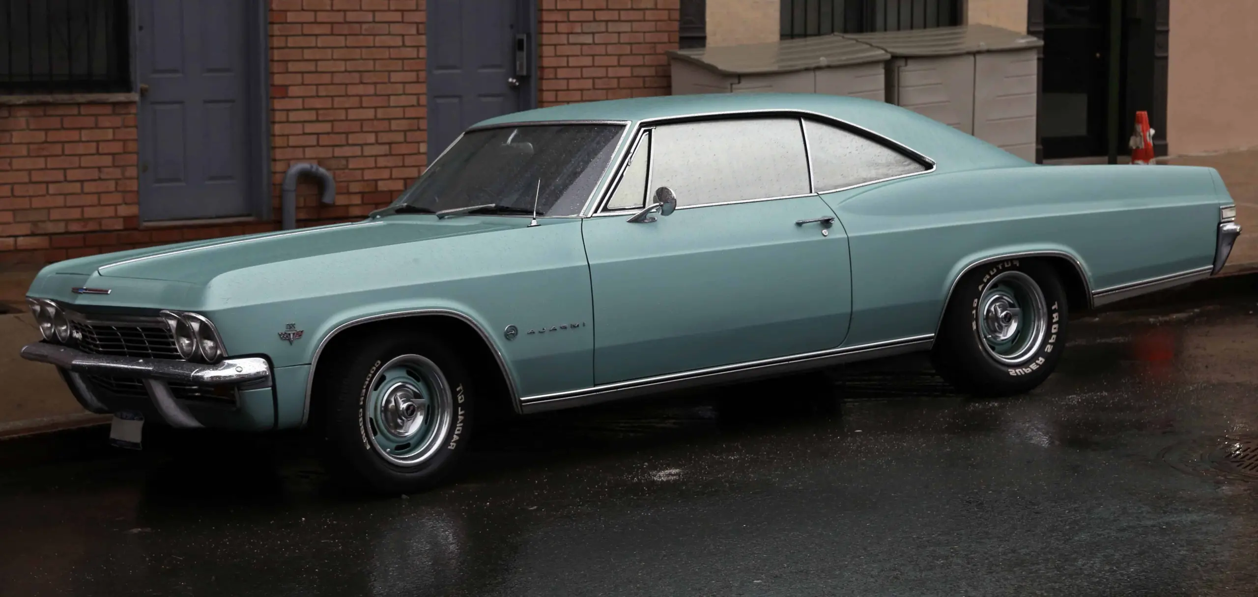 A light blue vintage Chevrolet Impala, reminiscent of stories found in prison writing, is parked on a wet street beside brick and stucco buildings, with water droplets visible on the car and pavement.