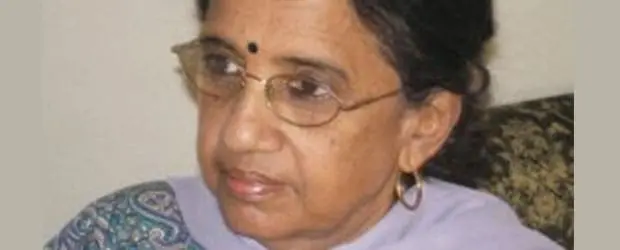 An older woman with glasses, a small black bindi on her forehead, gold earrings, and wearing a patterned shawl looks to the side with a neutral expression.