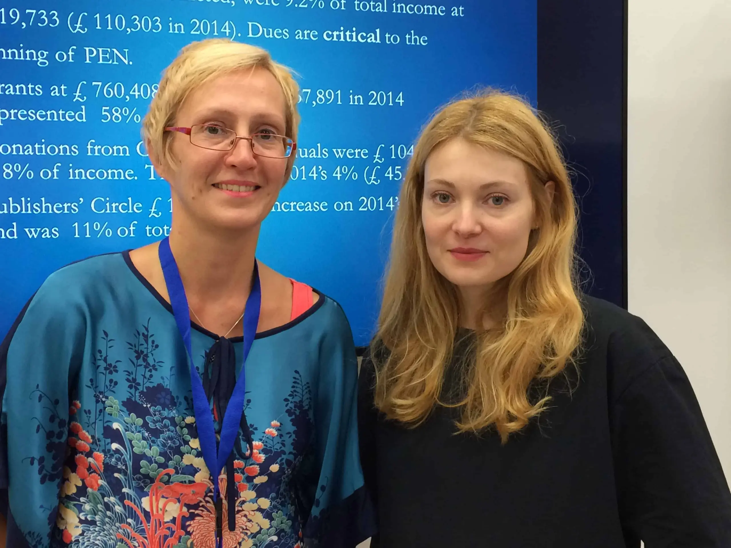 Two women stand side by side and smile at the camera. One wears glasses, a floral top, and a blue lanyard; the other has long blonde hair and a black shirt. A presentation slide with text is visible in the background.