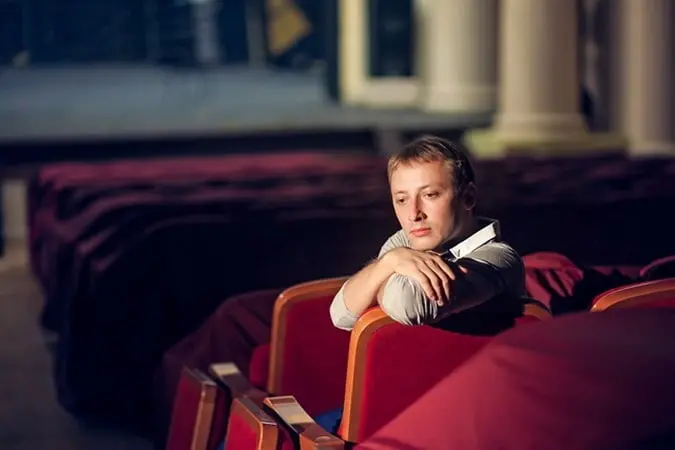 A person sits alone in a theater with red seats, resting their chin on their arms and looking thoughtful or contemplative. The setting appears quiet and empty.