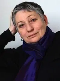 A person with short gray hair leans their head on one hand and looks at the camera. They are wearing a dark top and a deep blue scarf, with a neutral background behind them.