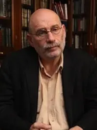 A middle-aged man with glasses, a bald head, and a gray beard sits in front of bookshelves, wearing a dark jacket over a light-colored shirt, looking thoughtfully at the camera.