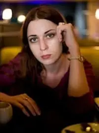 A woman with long brown hair sits at a table in a dimly lit setting, resting her head on one hand and looking directly at the camera with a neutral expression. A cup is visible on the table.