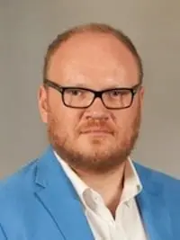 A man with short reddish hair and a beard wears black glasses, a white collared shirt, and a light blue blazer. He is looking at the camera with a neutral expression against a plain background.