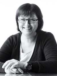 A woman with short hair and glasses is smiling and sitting at a table. She is wearing a dark cardigan over a light top. The image is in black and white.