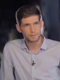 A young man with short brown hair, wearing a light blue button-up shirt with a microphone clipped to it, looks slightly to the side in a dimly lit setting.