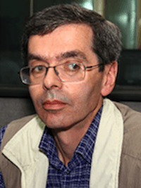 A middle-aged man with short dark hair, glasses, and a neutral expression is wearing a blue plaid shirt and a beige vest, sitting indoors.