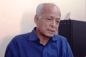 An older man with short gray hair, wearing a blue shirt, sits indoors against a light-colored wall, looking slightly downward with a thoughtful expression.