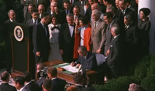 A man sits at a desk signing a document outdoors, surrounded by a large group of men and women watching. The U.S. presidential seal is visible on a podium nearby.