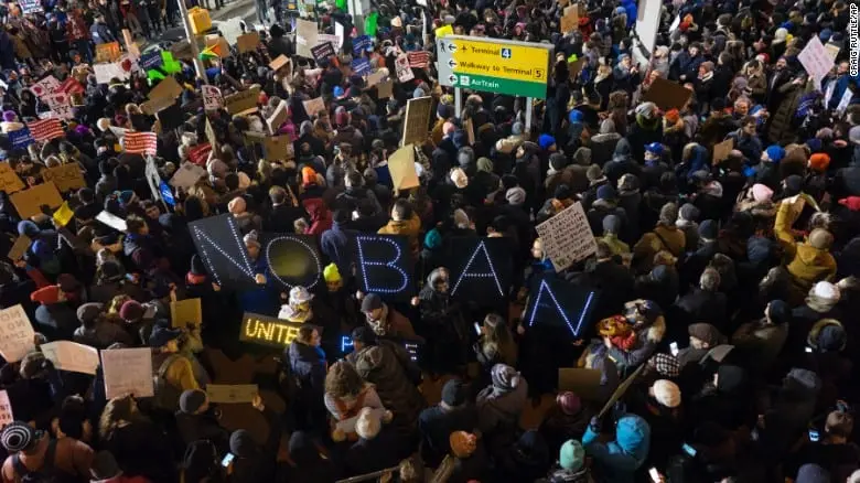 A large crowd of protesters holds signs and banners, including a lit-up sign that reads “NO BAN,” during a demonstration at an airport terminal. People are packed closely together, some waving small American flags.