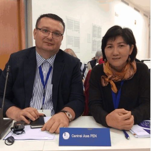 Two people sit side by side at a conference table with a sign reading Central Asia PEN in front of them. Both are wearing lanyards and formal attire, looking at the camera and smiling slightly.