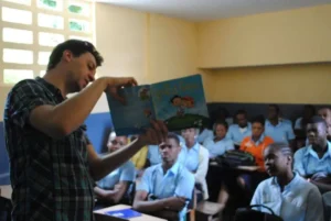 A man stands in front of a classroom, holding up and reading a colorful childrens book to a group of attentive students in school uniforms seated at desks.