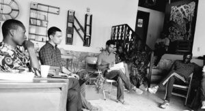 A group of six people sits in a relaxed, indoor setting, engaged in discussion. Some hold papers, one has a cup, and bookshelves are visible in the background. The image is in black and white.
