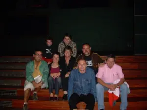 A group of eight adults sits on wooden steps, posing for a photo in a dimly lit setting. Four people sit on the top step and four on the lower step, all smiling at the camera.