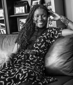 A smiling woman with long, braided hair sits on a couch, leaning against the armrest. She wears a patterned dress and bracelets. Framed photos are visible on shelves behind her. The image of Nicole Dennis-Benn is in black and white.